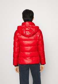 Czerwona kurtka puchowa o błyszczącym wykończeniu, pikowanym wzorze i kapturze. Posiada dopasowane mankiety i sięga talii, zestawiona z ciemnymi spodniami.