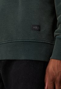 Felpa verde scuro con polsini e orlo a costine, con un'etichetta nera con la scritta "Calvin Klein Jeans" attaccata sul lato inferiore.