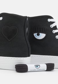 Czarne sneakersy za kostkę z białymi podeszwami, jeden but ozdobiony czarnym sercem, drugi niebieskim okiem z rzęsami, do tego pasujące czarne sznurowadła.