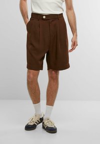 Braune, plissierte Shorts mit weitem Bein, ausgestattet mit Seitentaschen und einem Knopfverschluss vorne. Getragen mit weißen Socken und dunklen Sneakern.