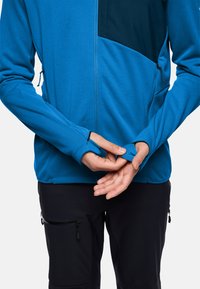 Giacca blue con zip, caratterizzata da una tasca sul petto di colore blu scuro, maniche aderenti e una texture morbida, abbinata a pantaloni neri con tasche con zip.