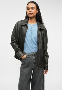 Καφέ δερμάτινο τζάκετ με φερμουάρ, ψηλό γιακά και ελαφριά γυαλάδα, φορεμένο πάνω από μπλε κάρντιγαν και γκρίζα φαρδιά τζιν.