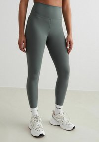 Hoge taille, aansluitende leggings in gedempt groen, gemaakt van gladde, rekbare stof. Gedragen met witte sokken en chunky witte sneakers.