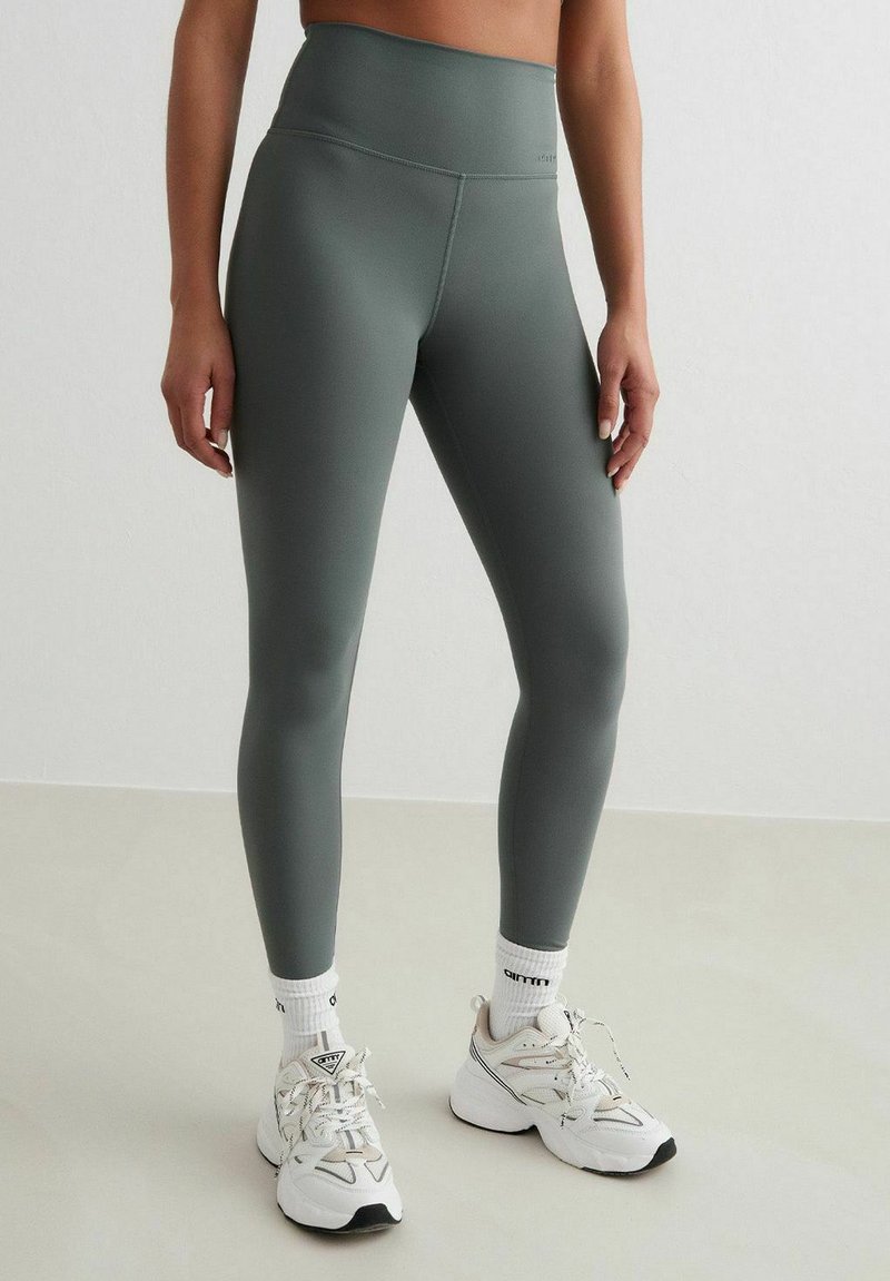 Hoge taille, aansluitende leggings in gedempt groen, gemaakt van gladde, rekbare stof. Gedragen met witte sokken en chunky witte sneakers.