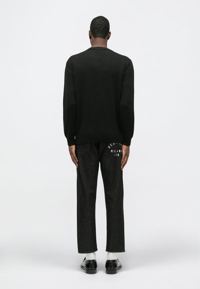 Vyras stovi atsigręžęs, vilkintis juodą megztinį, juodas džinsus su baltu užrašu ant nugaros, baltas kojines ir juodus elegantiškus batus ant vienodo fondo.