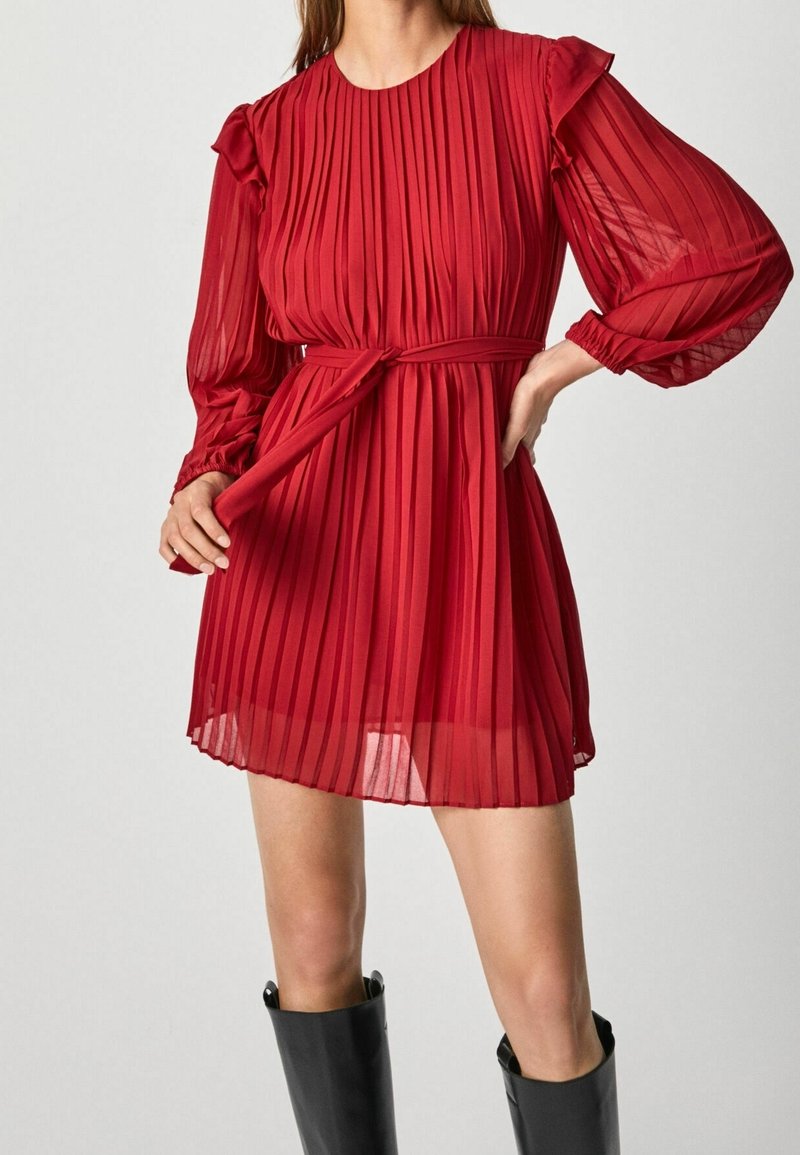 Robe longue rouge plissée à manches longues avec des volants aux épaules et une ceinture à nouer à la taille, portée avec des bottes noires montant jusqu'aux genoux, modèle debout, une main sur la hanche.
