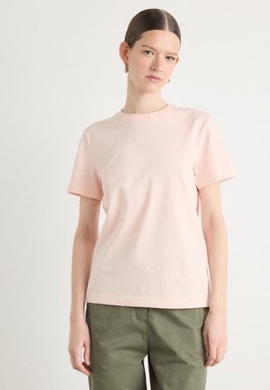 Lichtroze katoenen T-shirt met korte mouwen en een ronde halslijn. Bevat subtiele branding op de borst. Gepaarde met olijfgroene broeken.