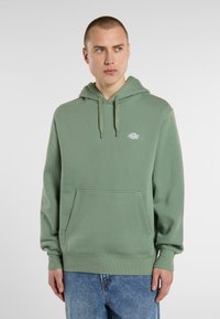 Manlig modell i en grön hoodie med ficka fram, parat med blå jeans, står framför en ljusgrå bakgrund.