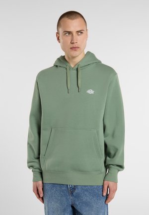 Mannelijke model in een groene sweatshirt met capuchon en een voorzak, gecombineerd met blauwe jeans, staat tegen een lichtgrijze achtergrond.