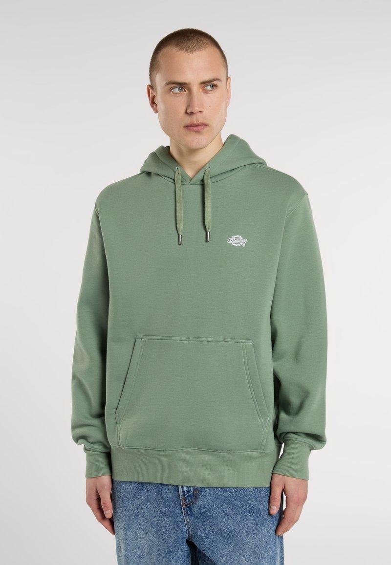 Manlig modell i en grön hoodie med ficka fram, parat med blå jeans, står framför en ljusgrå bakgrund.