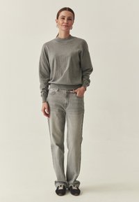 Pull en maille gris avec un col montant, coupe décontractée ; jean en denim gris clair ; baskets noires avec lacets blancs ; fond neutre.