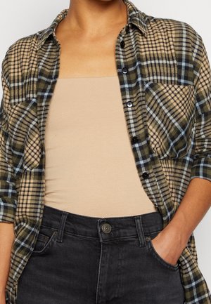 Person iført en olivengrøn og beige ternet skjorte over en beige tanktop og sorte jeans med den ene hånd i lommen.