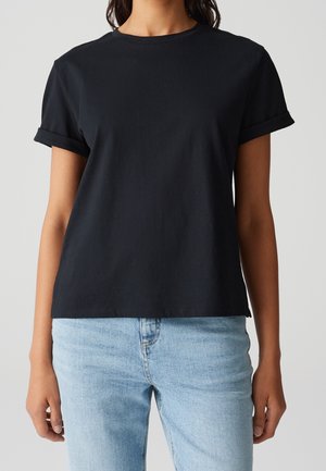 Femme portant un t-shirt noir uni à manches courtes retroussées et un jean taille haute bleu clair, se tenant devant un fond uni.