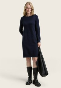 Marineblaues Strickkleid mit langen Ärmeln, gerippten Bündchen und Saum. Kombiniert mit knielangen schwarzen Gummistiefeln und einer großen schwarzen Tragetasche.