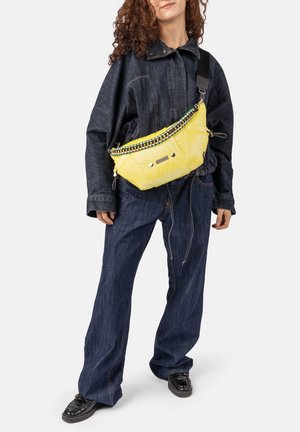 Personne portant une veste et un pantalon en denim foncé avec des chaussures noires, portant un grand sac bandoulière jaune vif avec une sangle tressée colorée.