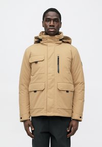 Mann trägt eine beige Kapuzen-Winterjacke mit mehreren Taschen und schwarze Hose, steht vor einem schlichten weißen Hintergrund.