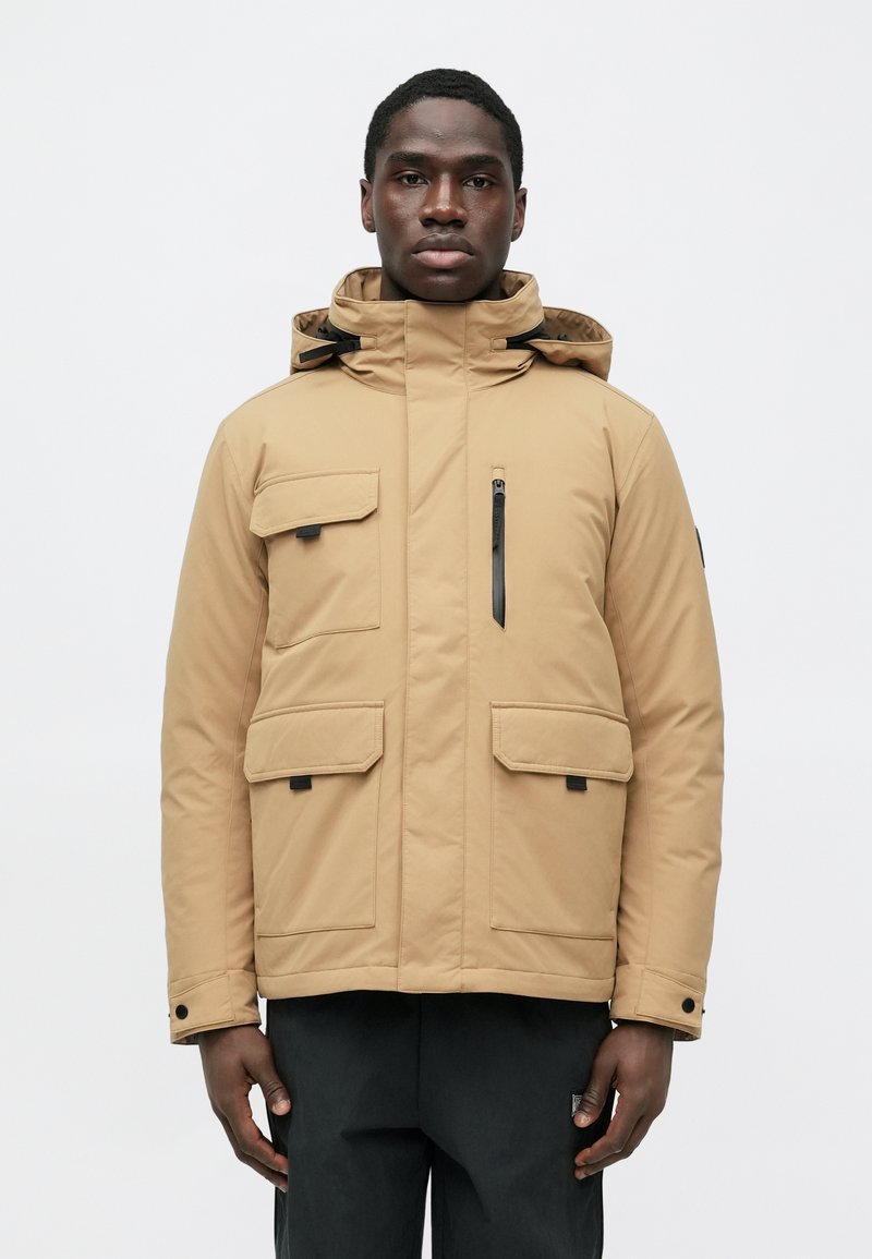 Mann trägt eine beige Kapuzen-Winterjacke mit mehreren Taschen und schwarze Hose, steht vor einem schlichten weißen Hintergrund.