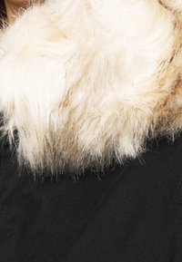 Svart jacka med en stor, mjuk, vit fuskpälskrage. Texturen är fluffig med långa fibrer, vilket kontrasterar med det släta tyget på jackan.