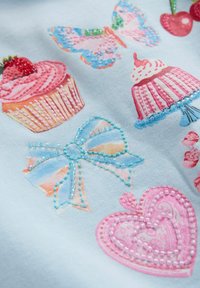 Tissu bleu clair avec des imprimés colorés de cupcakes, papillons, cerises et cœurs, agrémenté de détails en perles et de broderies.