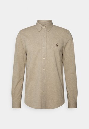 Beige langärmelig overhemd met knopen, een button-down kraag en een klein donker geborduurd logo op de linkerborst.