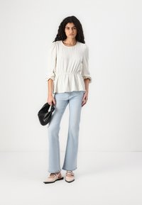 Blouse blanche à rayures avec taille froncée, jeans évasés bleu clair et chaussures à talons roses. Le modèle tient un petit sac à main noir.