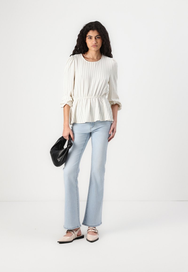 Blusa blanca a rayas con cintura fruncida, jeans de mezclilla azul claro acampanados y zapatos de punta rosados. La modelo sostiene un pequeño bolso negro.