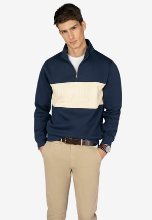 Sudadera navy con cuello de media cremallera, panel en crema en el pecho con el logo "HARPER & Neyer" en blanco, y puños acanalados, combinada con pantalones beige.
