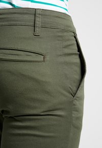 Spodnie w kolorze oliwkowej zieleni, wykonane z tkaniny z mieszanki bawełny, z tylną kieszenią i subtelnymi detalami szwów, o gładkiej fakturze.