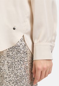 Cremefarbige Seidenbluse mit Knopfdetails und abgerundetem Saum, kombiniert mit einem Rock aus goldenen Pailletten. Der Ärmel der Bluse verfügt über eine glänzende Knopfakzentuierung.