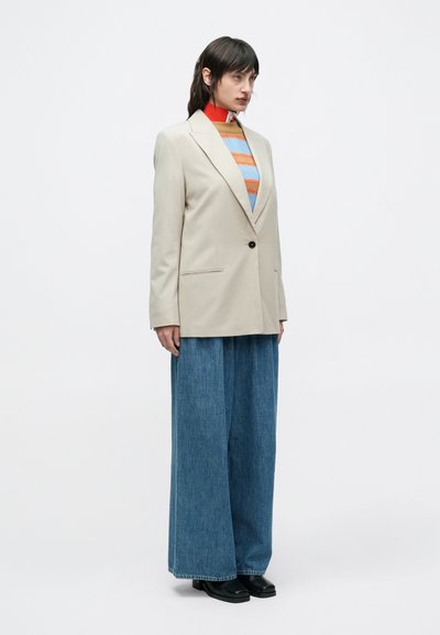 Μπεζ υπερμεγέθη μπλέιζερ με ένα μαύρο κουμπί, φορεμένο πάνω από πολύχρωμο ριγέ πουλόβερ, συνδυασμένο με παντελόνι τζιν με φαρδιές πόρπες και μαύρα παπούτσια.