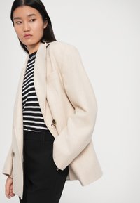 Woman with long dark hair wearing a beige blazer, black and white striped top, and black pants, looking to the side with one hand in pocket.