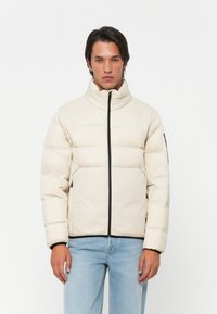 Un jeune homme aux cheveux raides et foncés porte une veste matelassée beige avec une fermeture éclair noire et un jean bleu clair, se tenant devant un fond blanc.