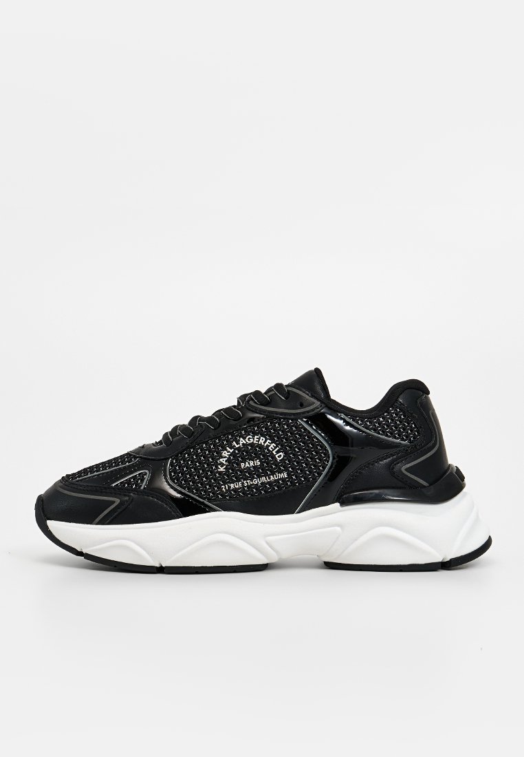 Zapatillas deportivas negras con parte superior de malla y acentos brillantes, suela gruesa blanca y cierre con cordones. Logo visible en el lateral.