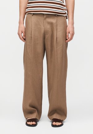 Osoba w luźnych beżowych spodniach, paskowanej bluzce bez rękawów i brązowych sandałach z odkrytymi palcami, stoi na jednolitym białym tle.