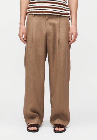 Personne portant un pantalon beige ample, un haut rayé sans manches et des sandales marron ouvertes aux orteils, debout devant un fond blanc uni.
