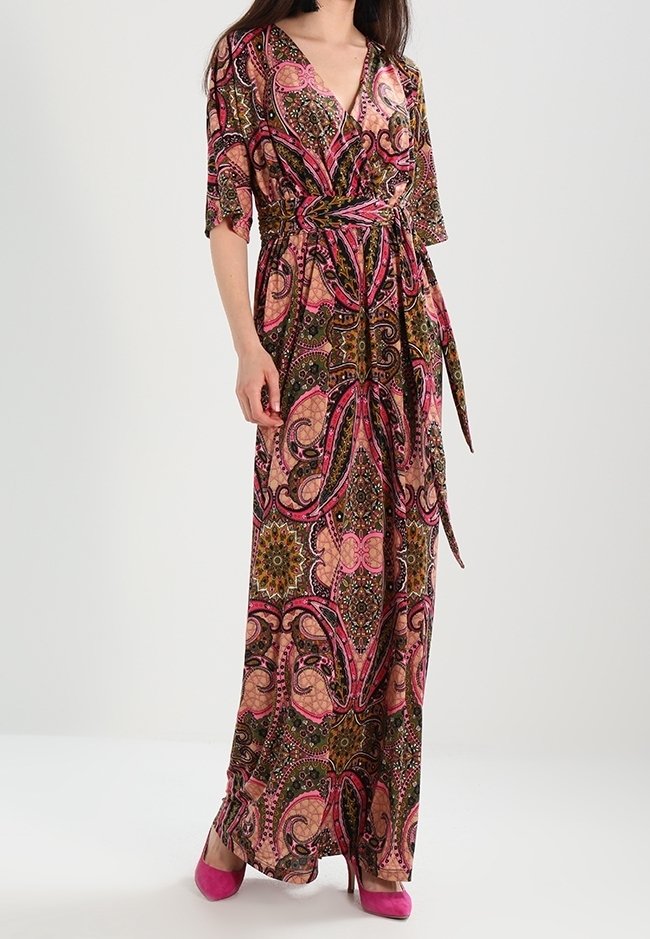 Robe maxi à motif cachemire coloré avec des tons roses, verts et bruns, ceinturée à la taille avec des manches courtes. Associée à des chaussures roses.