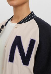 Veste beige clair avec manches bleu marine, présentant un grand "N" texturé foncé sur la poitrine, des boutons-pression dorés et un col côtelé rayé.