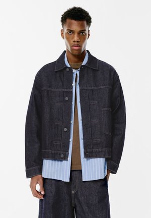 Mørkeblå denimjakke med en klassisk pasform, knaplukning foran og to brystlommer. Båret over en lyseblå stribet skjorte.