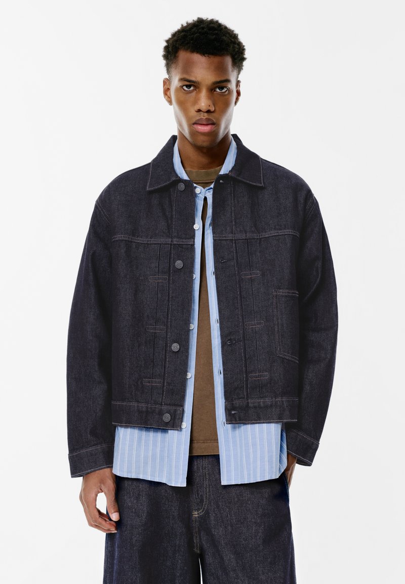 Tamno plava traper jakna klasičnog kroja, s dugmadi sprijeda i dva džepa na prsima. Nosena preko svjetloplavog prugastog košulje.