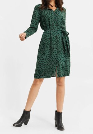 Frau trägt ein grünes, langärmliges Kleid mit Gürtel und schwarzem abstraktem Muster sowie schwarze Knöchelstiefel, steht vor weißem Hintergrund.