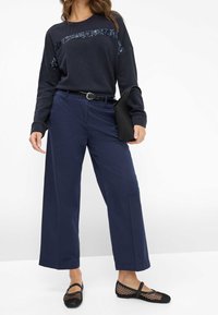 Dunkelblaues Sweatshirt mit einem Paillettenpanel, kombiniert mit weit geschnittenen Marineblauen Hosen und schwarzen Slip-On Schuhen aus Netzmaterial. Schwarzer Gürtel und Clutch sind ebenfalls enthalten.
