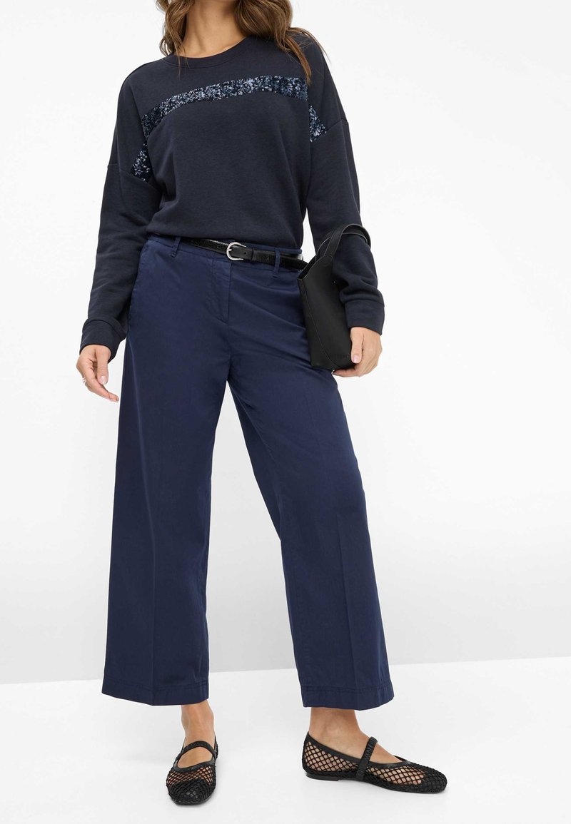 Dunkelblaues Sweatshirt mit einem Paillettenpanel, kombiniert mit weit geschnittenen Marineblauen Hosen und schwarzen Slip-On Schuhen aus Netzmaterial. Schwarzer Gürtel und Clutch sind ebenfalls enthalten.