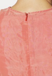 Růžové šaty s hladkým povrchem, s zadním zipem a červeným táhlem. Na látce je viditelný jemný vzor.