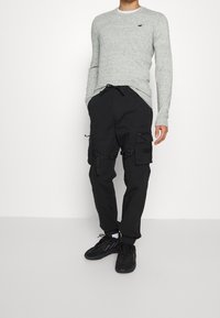 Šedé dlouhé tričko s texturou a malým logem, spárované s černými cargo kalhotami s několika kapsami a nastavitelnými popruhy. Černé tenisky.