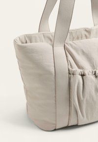 Bež torba za na ramo z napihnjeno teksturo, ki ima široke tkanine trakove in nabrano podrobnost na strani. Material se zdi mehak in lahek.