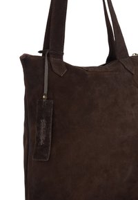 Bruine suède draagtas met dubbele schouderbanden, voorzien van een klein, aangehecht leren label en een gladde textuur. Minimalistisch ontwerp, zonder zichtbare sluitingen.