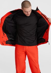 Veste orange avec doublure noire, dotée d'une fermeture éclair et de plusieurs poches, portée sur une chemise noire à manches longues et un pantalon orange assorti.