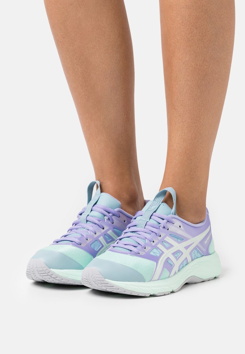 Jambes portant des chaussures de course pastel violet et vert menthe avec des semelles blanches, debout sur un fond clair uni.