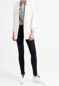 Light pink blazer with two buttons, paired with a camo-patterned shirt, black skinny jeans, and white sneakers with black accents.