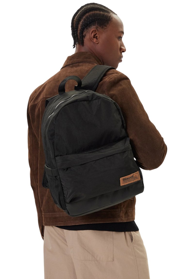 Uomo con giacca marrone e pantaloni beige che indossa uno zaino nero con tasca frontale e toppa in pelle sul retro.